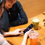 Two friends enjoy drinks at a cafe while looking at a stock market app on a smartphone.