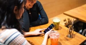 Two friends enjoy drinks at a cafe while looking at a stock market app on a smartphone.