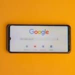 Smartphone displaying Google search page on a vibrant yellow background.
