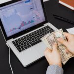 Person counting dollar bills with financial graphs on a laptop screen, symbolizing financial analysis and planning.