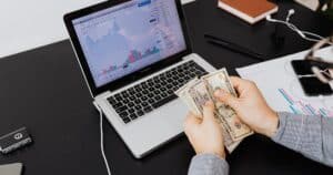 Person counting dollar bills with financial graphs on a laptop screen, symbolizing financial analysis and planning.